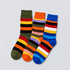 Three pairs of striped crew socks arranged overlapping on a light gray background. The left pair is olive green with red, yellow, black, and orange stripes; the middle pair is orange with purple, brown, pink, beige, and black stripes; the right pair is blue with red, white, yellow, black, and light blue stripes.